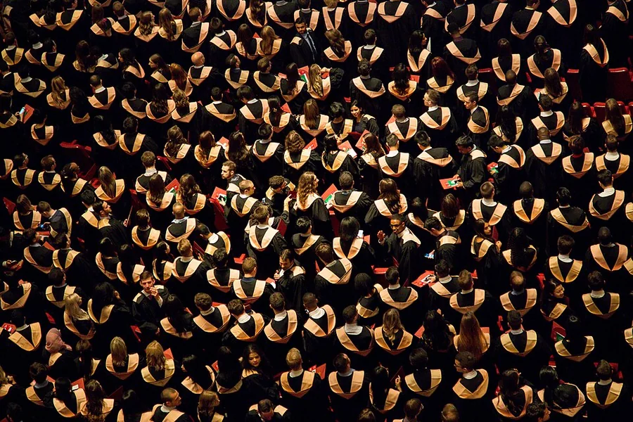 Felülről fényképezett nagy tömeg fekete talárban, arany színű gallérral egy diplomaosztó ünnepségen.