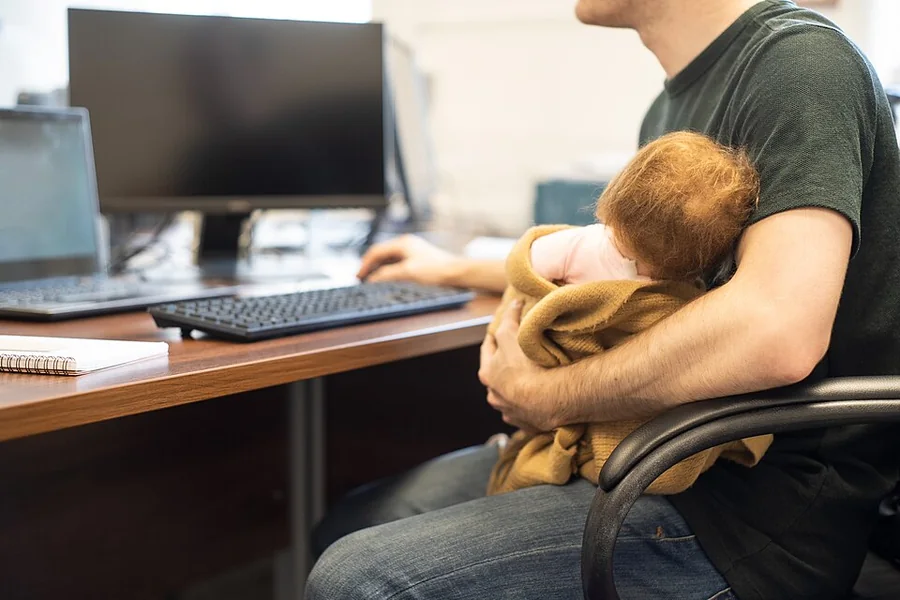 Egy férfi íróasztalnál ül, karjában takaróba bugyolált kisbabát tart, másik keze a billentyűzet mellett.