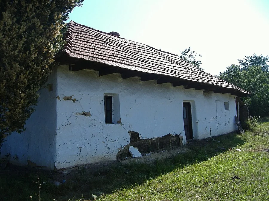 Régi, földszintes, fehérre meszelt parasztház, meredek tetővel, kis ablakokkal, a falon lepergett vakolat; előtte füves udvar, mellette fák.