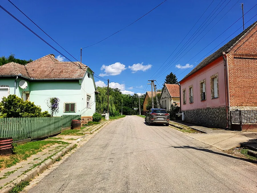 Magyar falu főutcája: üres aszfaltút, két oldalán alacsony, cseréptetős házak, kerítés, fák, egyetlen parkoló autó.