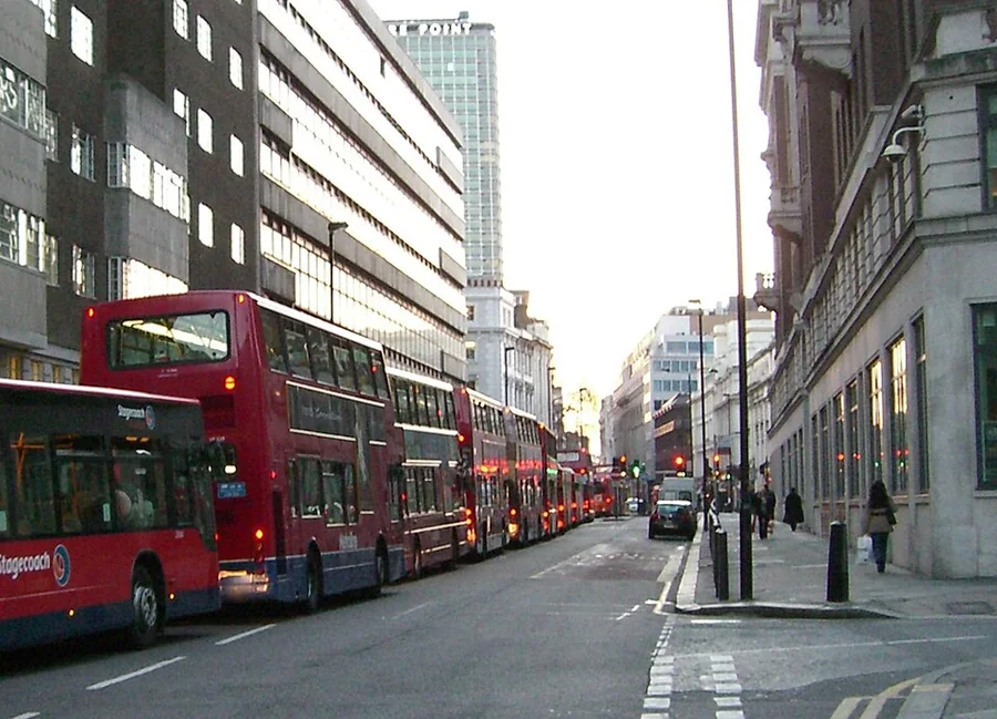 Londoni belvárosi utca, amelyen piros emeletes buszok hosszú sora áll egymás mögött; két oldalt magas épületek, a járdán néhány gyalogos.